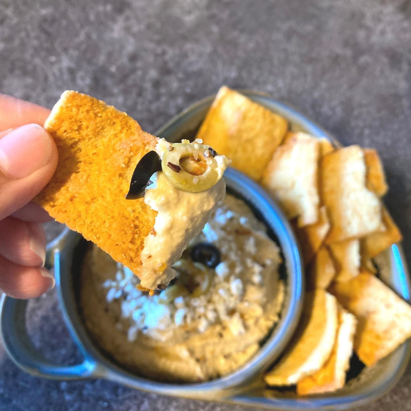 hand holding pita chip with scoop of everything hummus